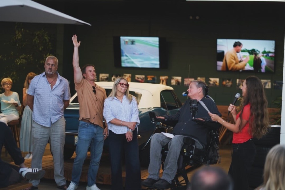 Sam Schmidt with family at the microphone during his birthday and book signing at the Petrol Club in San Clemente, a classic car and racing footage behind them
