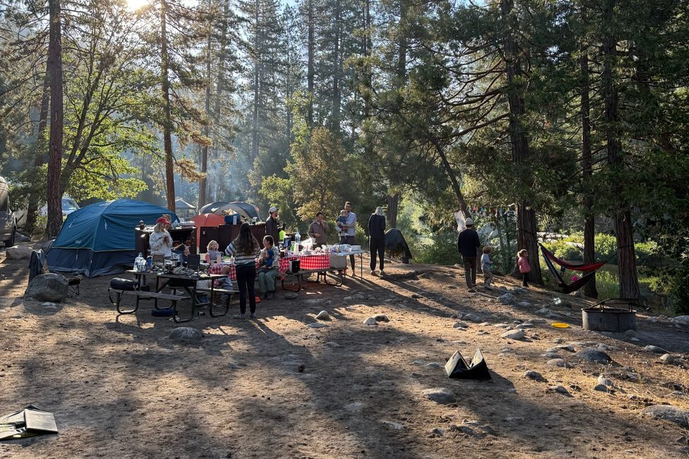 The campsite in the morning: two tents, picnic tables under a red gingham cloth, kids and parents standing around breakfast, a hammock strung between pines